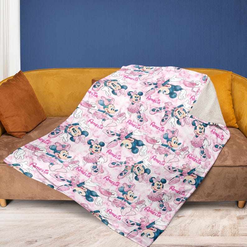 Puede incluir: Una manta rosa y blanca con un dise&ntilde;o de rat&oacute;n de dibujos animados con ropa de ballet. La manta est&aacute; sobre un sof&aacute; marr&oacute;n con un marco dorado. El nombre "Amelia" est&aacute; impreso en la manta.