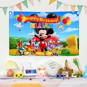 以下が含まれることがあります： ミッキーマウスと仲間たちが描かれた、カラフルな誕生日バナー。「Happy Birthday! ELIJAH」の文字入り。風船と青い空の背景。パーティーの飾り付けがバナーの前のテーブルに置かれています。