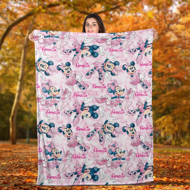 Puede incluir: Una manta rosa y blanca con un patr&oacute;n repetido de Minnie Mouse con atuendos de ballet y el nombre "Amelia". La manta tiene un fondo con efecto de m&aacute;rmol. Una persona sostiene la manta.