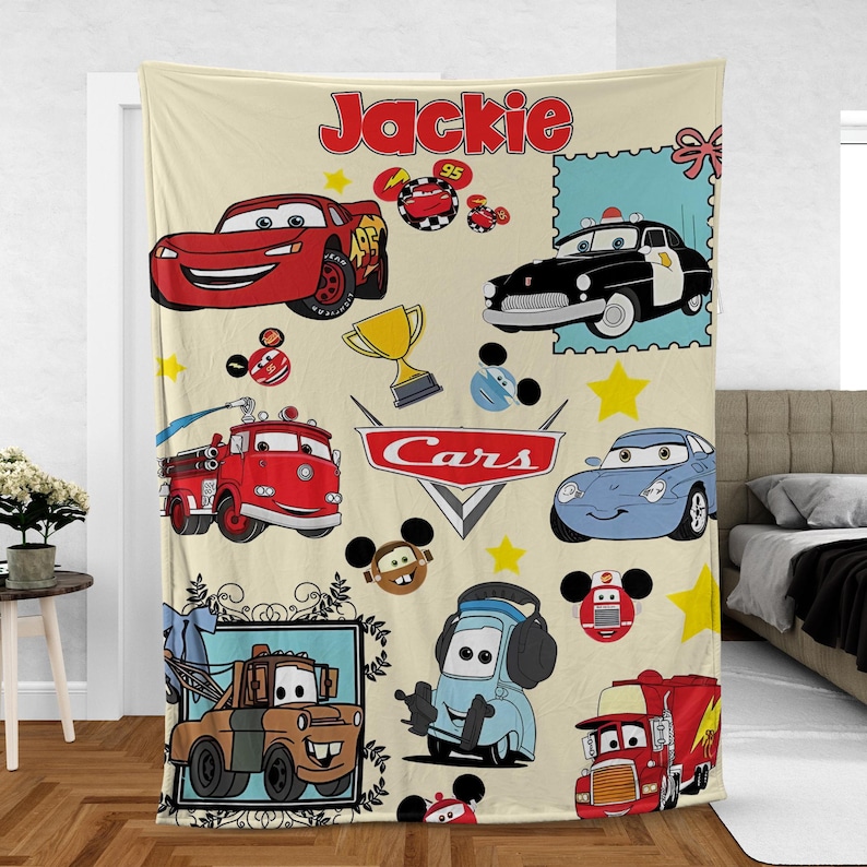 May include: A beige Cars-themed blanket featuring various cartoon car characters. The blanket includes the name "Jackie" in red script, along with stars, a trophy, and a Cars logo. The design is colorful and playful.