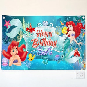 Puede incluir: Una pancarta rectangular con una escena submarina que muestra a la sirena de pelo rojo, un pez amarillo, un cangrejo rojo y una tortuga marina. El texto Happy Birthday Sofia 5 aparece en letras rojas, moradas y turquesas sobre un fondo azul.