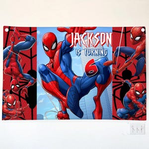 Pode incluir: Uma faixa retangular com o Homem-Aranha em v&aacute;rias poses de a&ccedil;&atilde;o contra um fundo vermelho e azul. O texto diz "JACKSON IS TURNING 6" em uma fonte estilizada. O design inclui um horizonte da cidade e padr&otilde;es de teia de aranha.