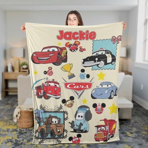 May include: A cream-colored blanket featuring various cartoon car characters, including a red race car, a blue car, and a fire truck. The blanket has the name "Jackie" in red script at the top and the "Cars" logo.