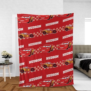 May include: Red fleece blanket featuring a repeating pattern of a cartoon race car with the number 95 and the name "HUDSON" in white. The blanket has a checkered flag design.