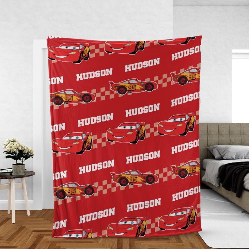 May include: Red fleece blanket featuring a repeating pattern of a cartoon race car with the number 95 and the name "HUDSON" in white. The blanket has a checkered flag design.