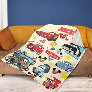 May include: A cream-colored Cars-themed blanket featuring various cartoon car characters, stars, and the word "Cars". The blanket has a sherpa lining and the name "Jacki" is printed on the top right corner.