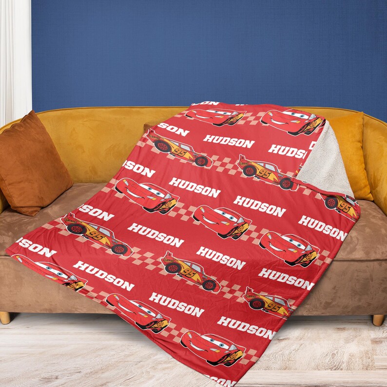 May include: A red blanket featuring a repeating pattern of a cartoon race car, the number 95, and the name "HUDSON" in white. The blanket is draped over a brown couch with a yellow pillow.