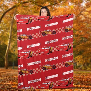 May include: Red blanket featuring a repeating pattern of cartoon race cars and the name "HUDSON" in white. The cars are red and yellow with the number "95" on them. The blanket is held up against a backdrop of fall foliage.