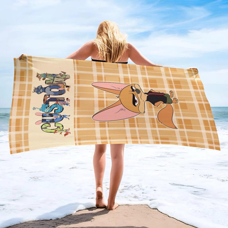 May include: A beach towel with a cartoon fox character and the word "CUSTOM" in colorful letters. The towel has a yellow plaid pattern and is held by a person on a beach.