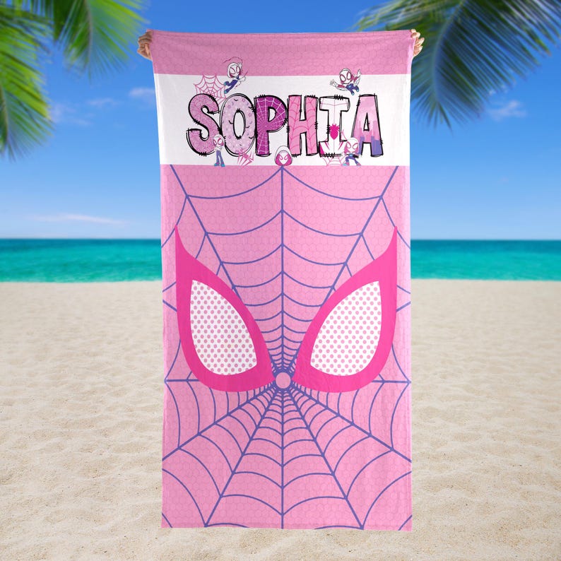 May include: Pink beach towel featuring a purple spiderweb design and large pink eye shapes. The name "SOPHIA" is printed in large letters across the top, with cartoon characters. The towel is held up against a beach background.
