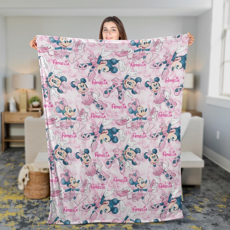 Puede incluir: Una manta rosa y blanca con un patr&oacute;n repetido de Minnie Mouse con atuendos de ballet. El dise&ntilde;o incluye el nombre "Amelia" en escritura rosa. La manta tiene un fondo con efecto de m&aacute;rmol. Un art&iacute;culo c&oacute;modo y decorativo para el hogar.