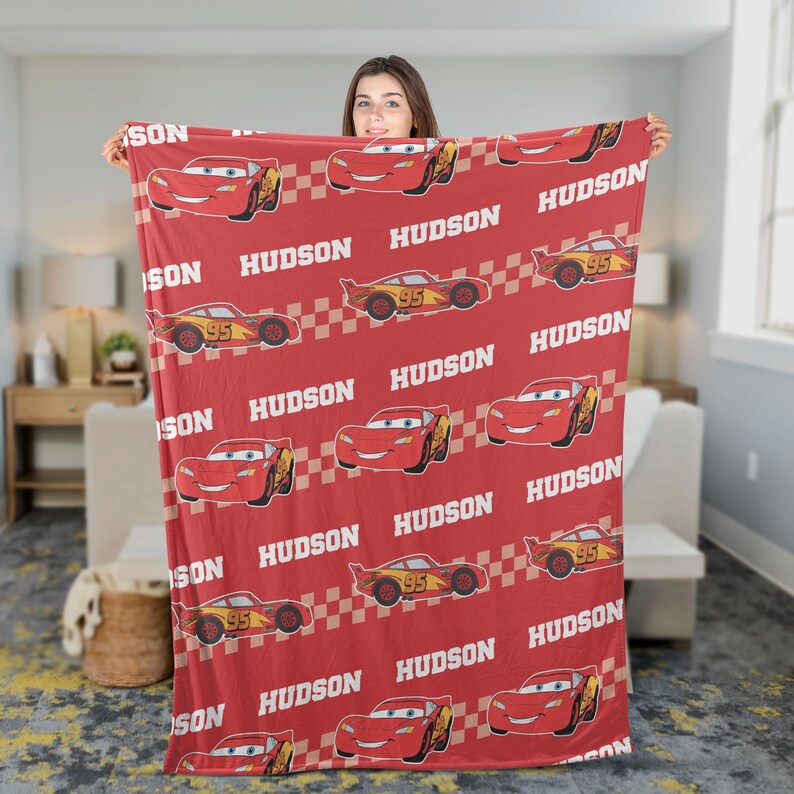 May include: Red blanket featuring a repeating pattern of a cartoon race car with the number 95, the name "HUDSON", and a checkered flag design. The blanket is held up by a person.