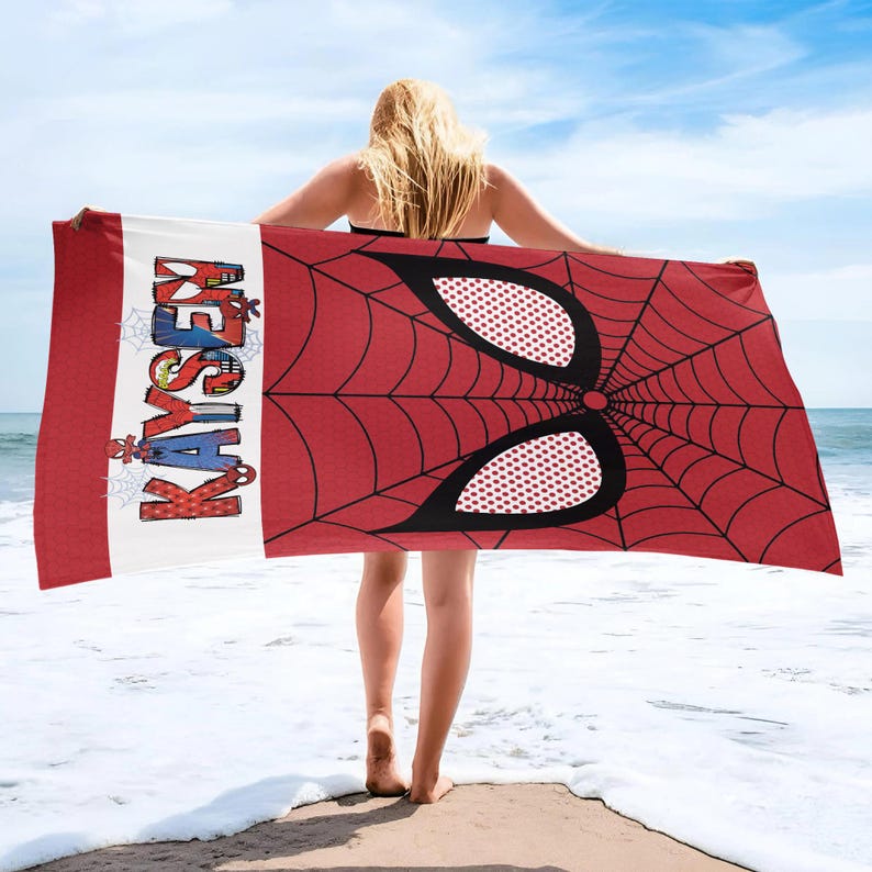 May include: A red and black beach towel featuring a Spider-Man mask design. The towel has a white stripe with the name "KAYSE" in colorful letters. The background shows a beach and ocean.