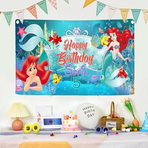 Puede incluir: Una pancarta de cumplea&ntilde;os rectangular con una escena submarina azul y personajes de La Sirenita, incluyendo a Ariel, Flounder y Sebasti&aacute;n. El centro muestra el texto Happy Birthday Sofia 5 en letras rojas y moradas.