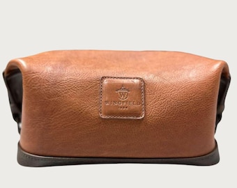 May include: A brown leather toiletry bag with a rectangular shape. The bag features a small, square patch with the "WINGFIELD 1934" logo. The bag has a dark brown base and a rounded top.