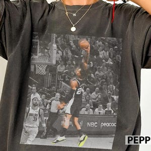 Pode incluir: Uma t-shirt cinzento-carvão com uma grande impressão gráfica a preto e branco de um jogador de basquetebol a fazer um afundanço, com outro jogador com a camisola Dallas 31, sobre um fundo de arena cheia com o texto NBC.