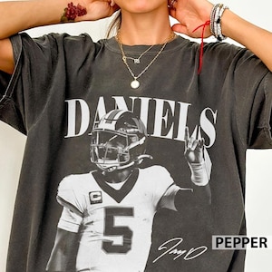 May include: A charcoal grey short-sleeve t-shirt featuring a large white graphic print of a football player wearing a helmet and a jersey with the number 5. The word DANIELS is printed in large white capital letters above the player graphic.