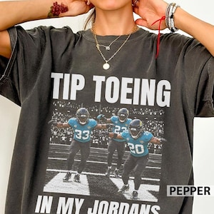 May include: A charcoal gray t-shirt featuring the white text TIP TOEING IN MY JORDANS. The graphic displays two football players in teal and white uniforms with numbers 33 and 20, running on a field during a game in a stadium.