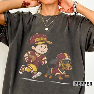 May include: A charcoal grey t-shirt featuring a graphic of a cartoon character wearing a burgundy and gold football uniform and a cap with the text WASHINGTON, running alongside a dachshund wearing a matching football helmet and jersey.