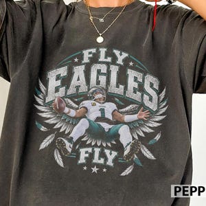 Può includere: Una t-shirt grigio antracite a maniche corte con la grafica di un giocatore di football in maglia bianca numero 1 che tiene un pallone. Il testo FLY EAGLES FLY è circondato da grandi ali bianche e stelle su sfondo circolare.