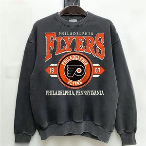 Könnte beinhalten: Ein anthrazitfarbenes Sweatshirt mit Rundhalsausschnitt und einem großen orange-weißen Grafikdruck mit der Aufschrift Philadelphia Flyers, dem Teamlogo, den Zahlen 19 und 67 sowie dem Text Philadelphia, Pennsylvania auf der Vorderseite.
