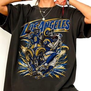 May include: A black short-sleeve t-shirt featuring a graphic print of two skeleton football players in blue and yellow uniforms with the text LOS ANGELES. A large ram head is centred below the players, surrounded by blue and yellow abstract shapes.