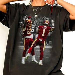 May include: A black short-sleeve t-shirt featuring a graphic print of two football players in burgundy and gold uniforms. One player wears the number 1 with the name WHITE on the back. The print includes two handwritten signatures and a blurred background.