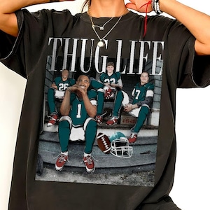 Peut inclure: Un t-shirt gris anthracite avec une impression graphique de style vintage portant le texte THUG LIFE en haut. L'image montre quatre joueurs de football en uniformes bleu sarcelle et blanc assis sur des marches en béton, avec un ballon et un casque.