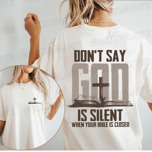 Puede incluir: Camiseta color crema con el texto "DON'T SAY GOD IS SILENT WHEN YOUR BIBLE IS CLOSED" en marrón oscuro y gris. El diseño incluye una cruz y un libro abierto. Una versión más pequeña del diseño está en la parte delantera.