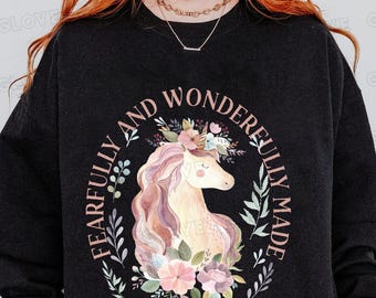 Op de afbeelding: Zwart sweatshirt met een roze en witte eenhoornillustratie omringd door bloemen en bladeren. De tekst "Fearfully and Wonderfully Made" staat in een cirkel rond de eenhoorn. De tekst "Psalm 139:14" staat onder de eenhoorn.
