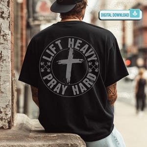 May include: A black short-sleeve t-shirt featuring a large circular graphic on the back. The design includes a distressed cross in the center, flanked by two small barbell icons, with the text LIFT HEAVY PRAY HARD printed around the circle.