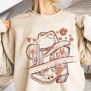 May include: A beige sweatshirt with a graphic design of a cowboy boot, a cactus, and a cowboy hat. The graphic is in shades of brown and pink. The text "I can do all things through Christ who gives me strength" and "Philippians 4:13" are included in the design.