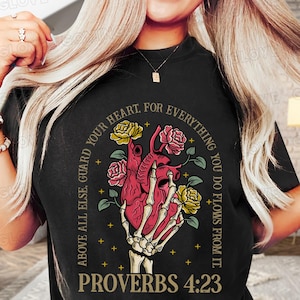 以下が含まれることがあります： 花柄の赤いハートを骨格の手が持っているグラフィックがプリントされた黒いTシャツ。グラフィックの周りに「ABOVE ALL ELSE, GUARD YOUR HEART. FOR EVERYTHING YOU DO FLOWS FROM IT.」というテキストが金色の円の中に書かれている。グラフィックの下に「PROVERBS 4:23」というテキストが書かれている。