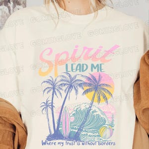 May include: A white t-shirt with a colorful graphic design featuring palm trees, a wave, and a surfboard. The text "Spirit Lead Me" is written in a colorful font. The scripture "Psalm 143:8-10" and the phrase "Where my trust is without borders" are also included in the design.