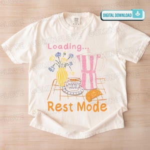 Op de afbeelding: Crèmekleurig T-shirt met een grillige illustratie. Het ontwerp toont een koffiepot, kopje, croissant en bloemen, met de woorden "Loading..." en "Rest Mode" in roze en oranje. Een digitaal downloadpictogram staat rechtsboven.