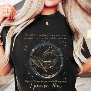 May include: Black t-shirt with a gold and white design featuring two whales swimming in a starry sky. The text "The LORD is my strength and my shield: my heart trusts in Him, and He helps me. Psalm 28:7" is printed on the shirt. The text "My heart leaps for joy, and with my song I praise Him" is also printed on the shirt.