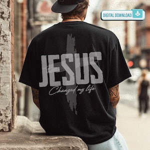 Może przedstawiać: Czarna koszulka z napisem "JESUS" dużymi, jasnoszarymi literami i szarym wzorem krzyża. Poniżej słowa "JESUS" znajduje się fraza "Changed my life" w kursywie. Koszulka jest noszona przez osobę.