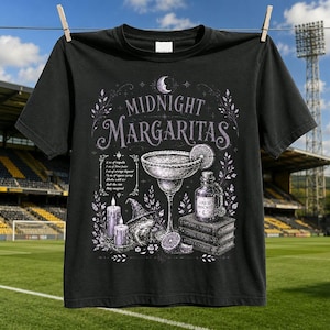 以下が含まれることがあります： 「MIDNIGHT MARGARITAS」の文字がプリントされた黒いTシャツ。デザインには、マルガリータグラス、魔女の帽子をかぶったカエル、2本のキャンドル、積み重ねられた本、レシピが描かれており、全体的に紫と白の色調でまとめられています。