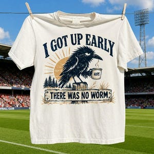 May include: A cream-coloured short-sleeve t-shirt featuring a graphic of a black crow perched on a wooden post, holding a mug labelled NOPE. The text reads I GOT UP EARLY and THERE WAS NO WORM, with a sun and pine tree silhouette background.
