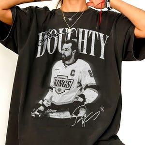 May include: A black t-shirt featuring a large white graphic of hockey player Drew Doughty in a Los Angeles Kings jersey. The text DOUGHTY is printed in a distressed white font above the player, with a signature and the number 8 below.
