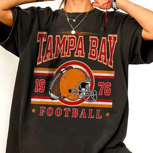 May include: A black short-sleeve t-shirt featuring a distressed graphic print that reads TAMPA BAY in red letters above an orange football helmet. The design includes the numbers 19 and 76 on either side, with the word FOOTBALL printed below.