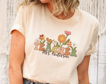 Pode incluir: Uma camiseta bege claro com um gráfico da palavra "TEACHER" em letras coloridas com flores e um rosto sorridente. O texto "Mrs. Anderson" está abaixo da palavra "TEACHER".