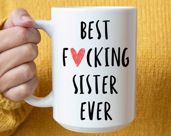 Könnte beinhalten: Weißer Keramikbecher mit dem Text "BEST FUCKING SISTER EVER" in Schwarz, mit einem roten Herz, das das "O" in "FUCKING" ersetzt. Der Becher wird von einer Person gehalten, die einen gelben Pullover trägt.
