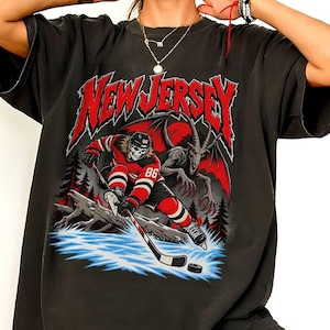 Könnte beinhalten: Ein schwarzes T-Shirt mit einem Grafikdruck und dem Text NEW JERSEY in Rot. Das Design zeigt einen skelettierten Eishockeyspieler in einem rot-schwarzen Trikot mit der Nummer 86, der auf Eis vor einer geflügelten Kreatur in einem Wald läuft.