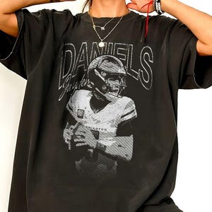 May include: A black short-sleeve t-shirt featuring a white graphic print of a football player in a helmet and jersey. The text "DANIELS" appears in large white letters above the player, with "Jayden" written in script below the name.