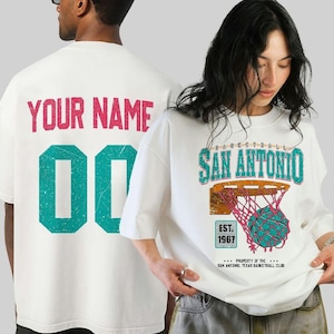 Pode incluir: T-shirts brancas com gráficos de basquete. Uma apresenta "YOUR NAME" e o número "00" em rosa e verde-azulado. A outra mostra "SAN ANTONIO" com um design de rede e bola de basquete, e "EST. 1967".