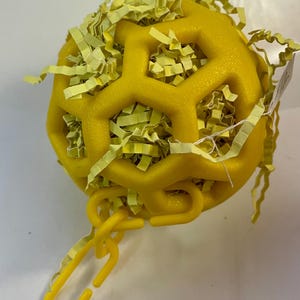 May include: Yellow plastic toy ball with a honeycomb design and a chain attached. The ball is filled with yellow shredded paper.