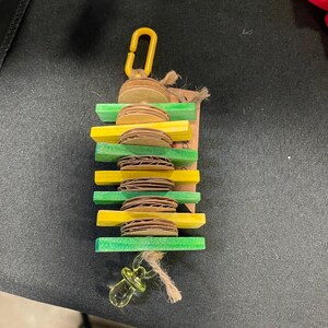 May include: A colorful bird toy featuring alternating yellow and green wooden blocks with cardboard discs. A yellow plastic clip and a clear pacifier-shaped charm are attached with twine. The toy is designed for birds.