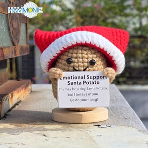May include: A crocheted Santa potato wearing a red and white Santa hat holds a sign that reads "Emotional Support Santa Potato. I'm may be a tiny Santa Potato, but I believe in you. Go do your thing!"
