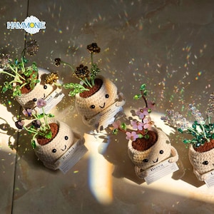 May include: Handmade crochet planters with smiling faces, each holding a name tag. The planters contain beaded flowers in various colors, including purple, pink, and brown. The planters are beige and sit on a light-colored surface.